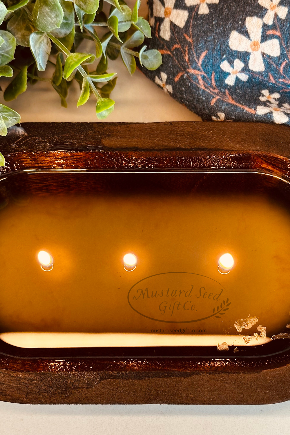 Candle in a decorative wooden holder with 'Mustard Seed Gift Co.' branding, surrounded by greenery.