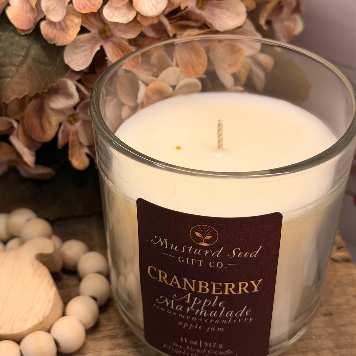 A glass jar candle with creamy white wax and a single wick, featuring a dark brown label that reads 'Mustard Seed Gift Co. Cranberry Apple Marmalade.