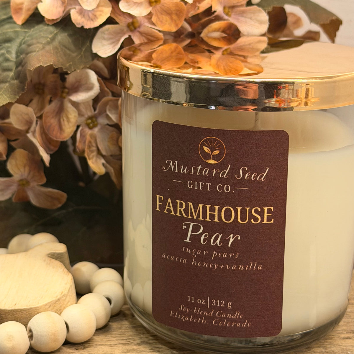 A Farmhouse Pear scented soy candle by Mustard Seed Gift Co. in a clear glass jar with a gold lid and a dark brown label, styled with dried flowers and wooden beads on a wooden surface.