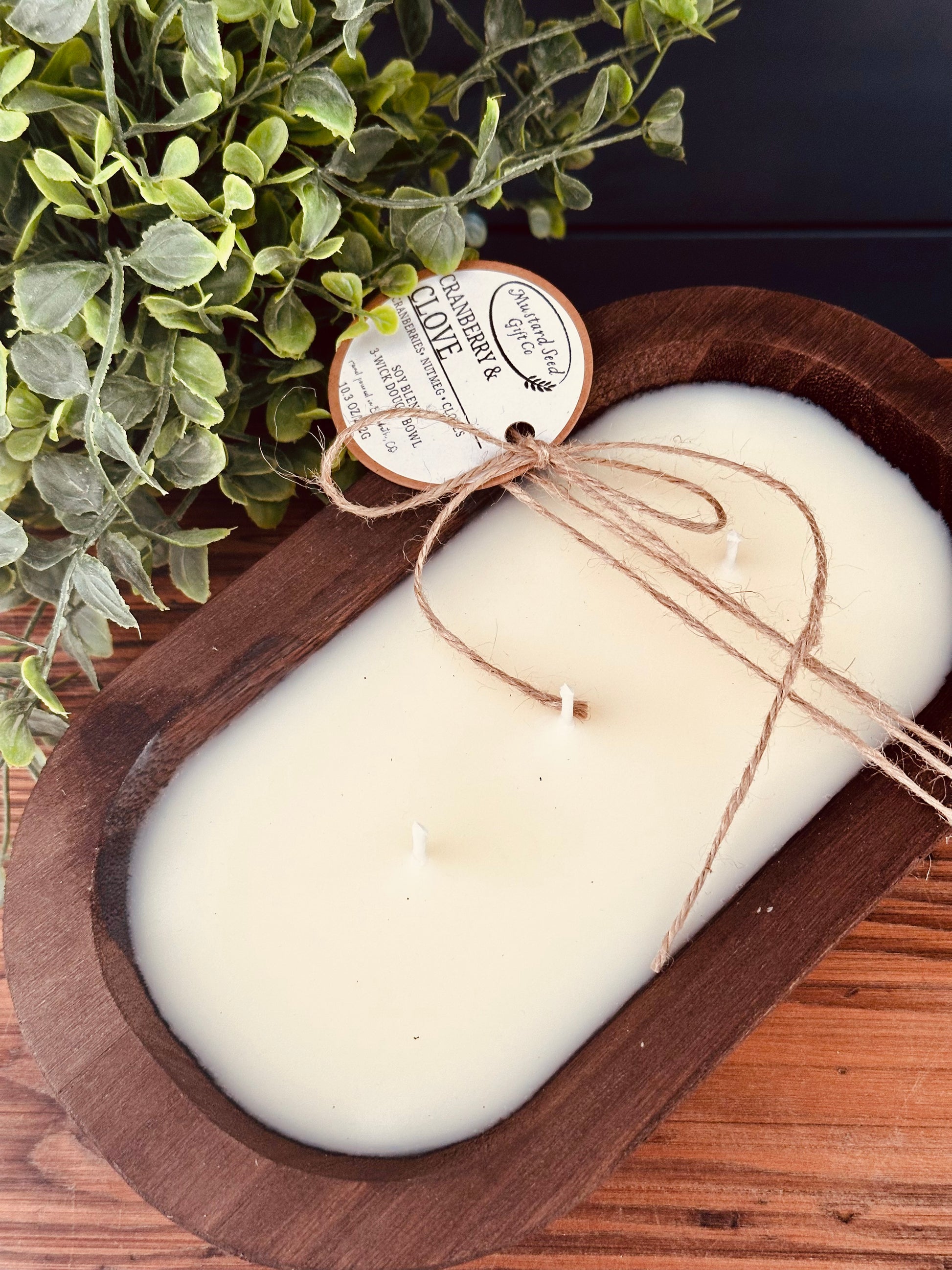 White candle in a wooden dish with a label, surrounded by greenery on a wooden surface.