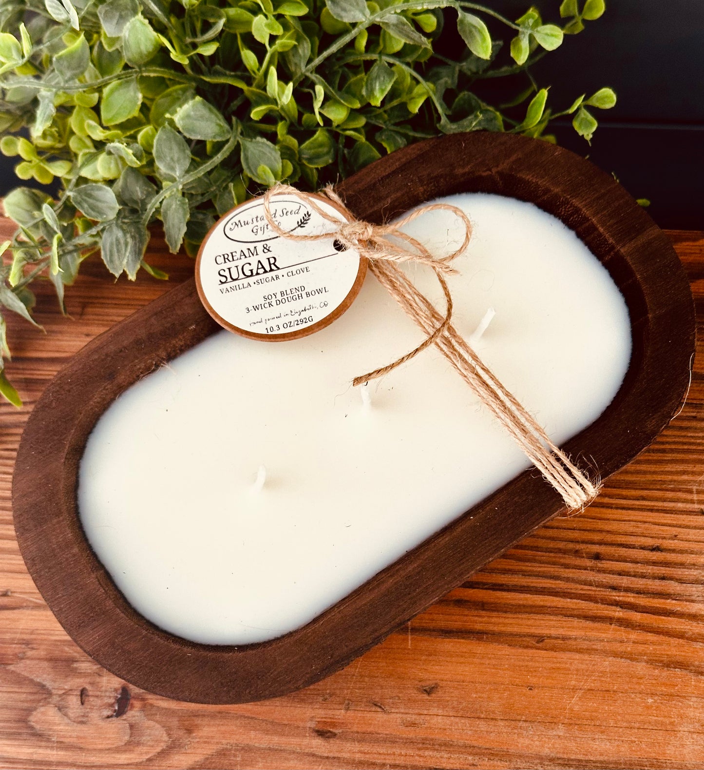 White candle in a wooden dish with a label, on a wooden surface with greenery in the background.