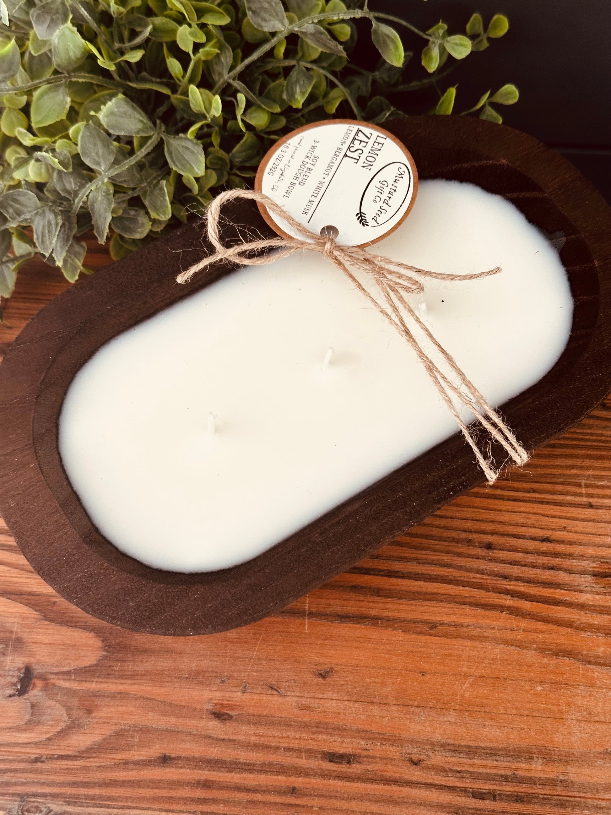 White candle in a wooden holder with a label, on a wooden surface with greenery in the background.
