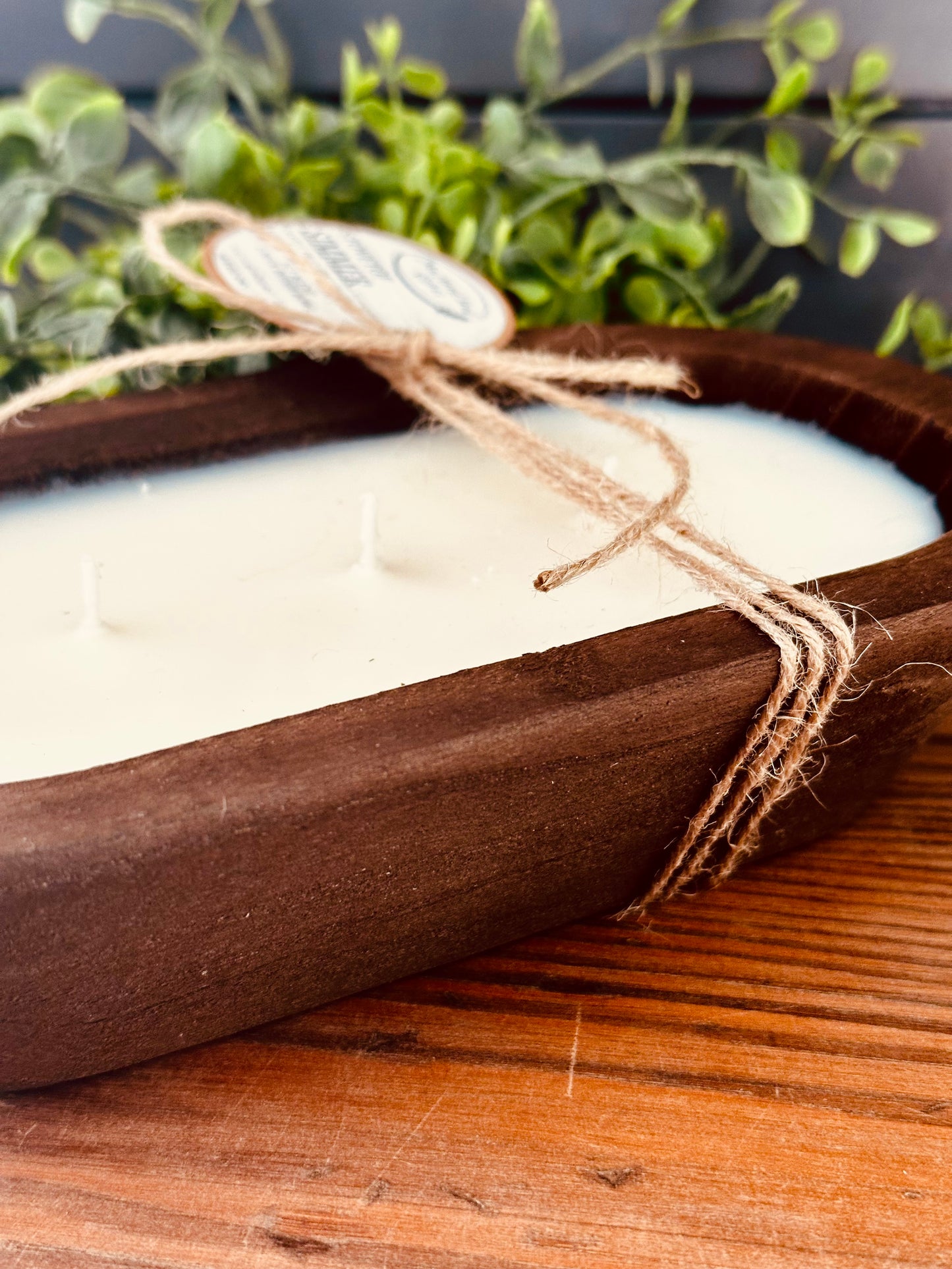 Candle in a wooden bowl with twine and a tag, on a wooden surface with greenery in the background.